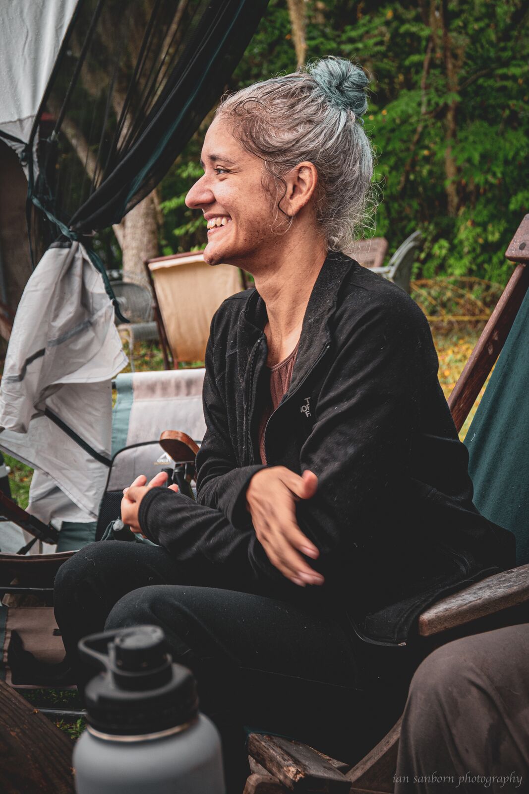 A person with grey hair in a topknot sits in a camp chair under a tarp shelter, laughing with their arms folded, a steel water bottle by their feet.
