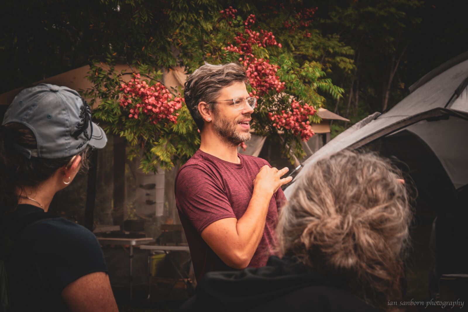 A man in a maroon shirt signs to two other people outside a tent, his hand raised mid-sign, a shrub full of red berries behind him.
