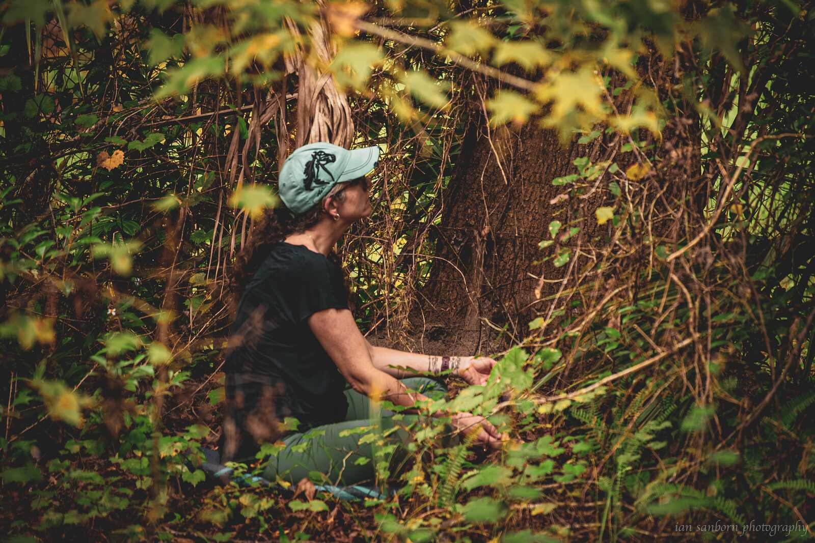 A person in a pale green cap sits low in a thicket of vines and autumn leaves, hands resting in the undergrowth, almost hidden by the foliage.