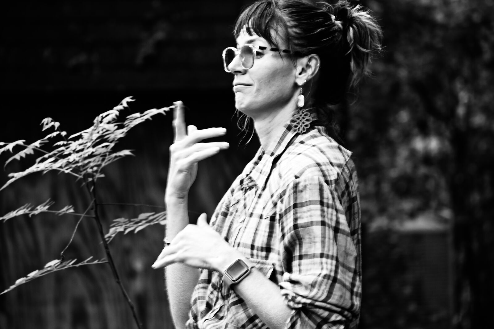 Black and white photograph of a person in round glasses and a plaid shirt signing beside a tall seed-headed grass stem.