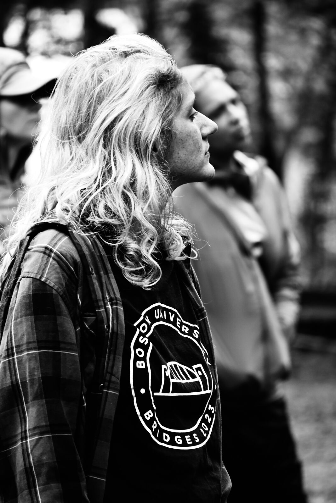 Black and white photograph of a person with long wavy hair in a plaid shirt, seen from behind in profile, standing among others in a group.