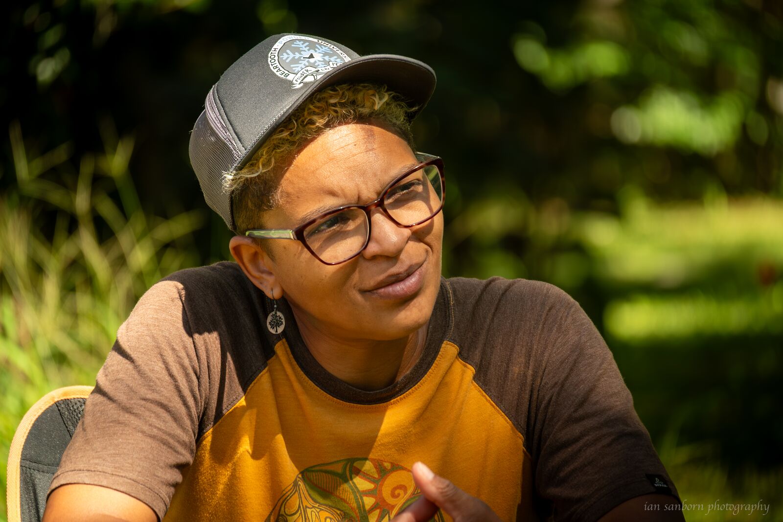 A person in a cap, glasses, and a yellow shirt sits outdoors mid-conversation, hands raised, sunlit greenery blurred behind them.