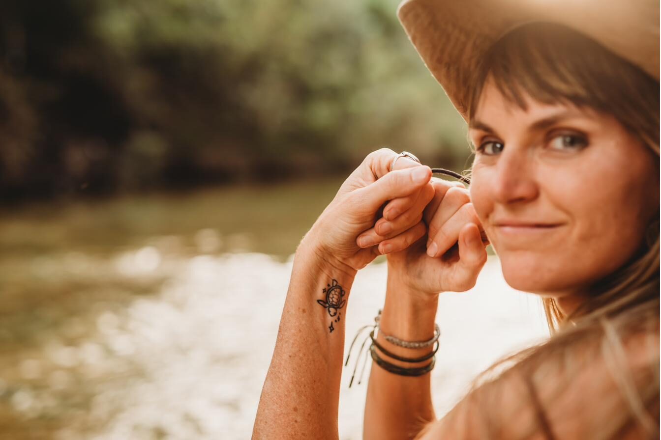 Summer Crider resting their hands against their cheek by a sunlit river, a small sea turtle tattoo on their forearm, looking calmly toward the camera.