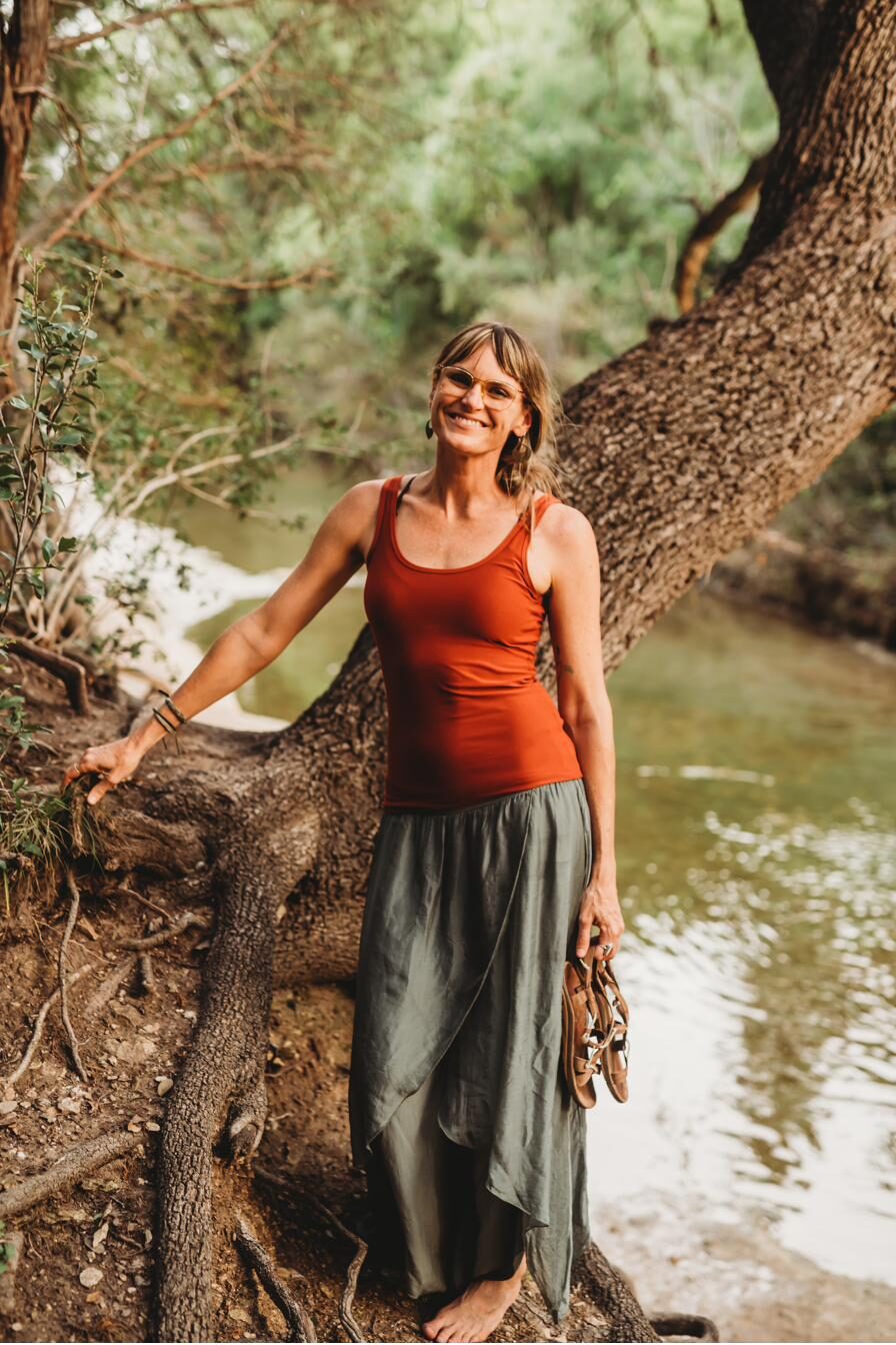 Summer Crider barefoot beside a creek, one hand on the trunk of a live oak that leans out over the green water, sandals in their other hand.